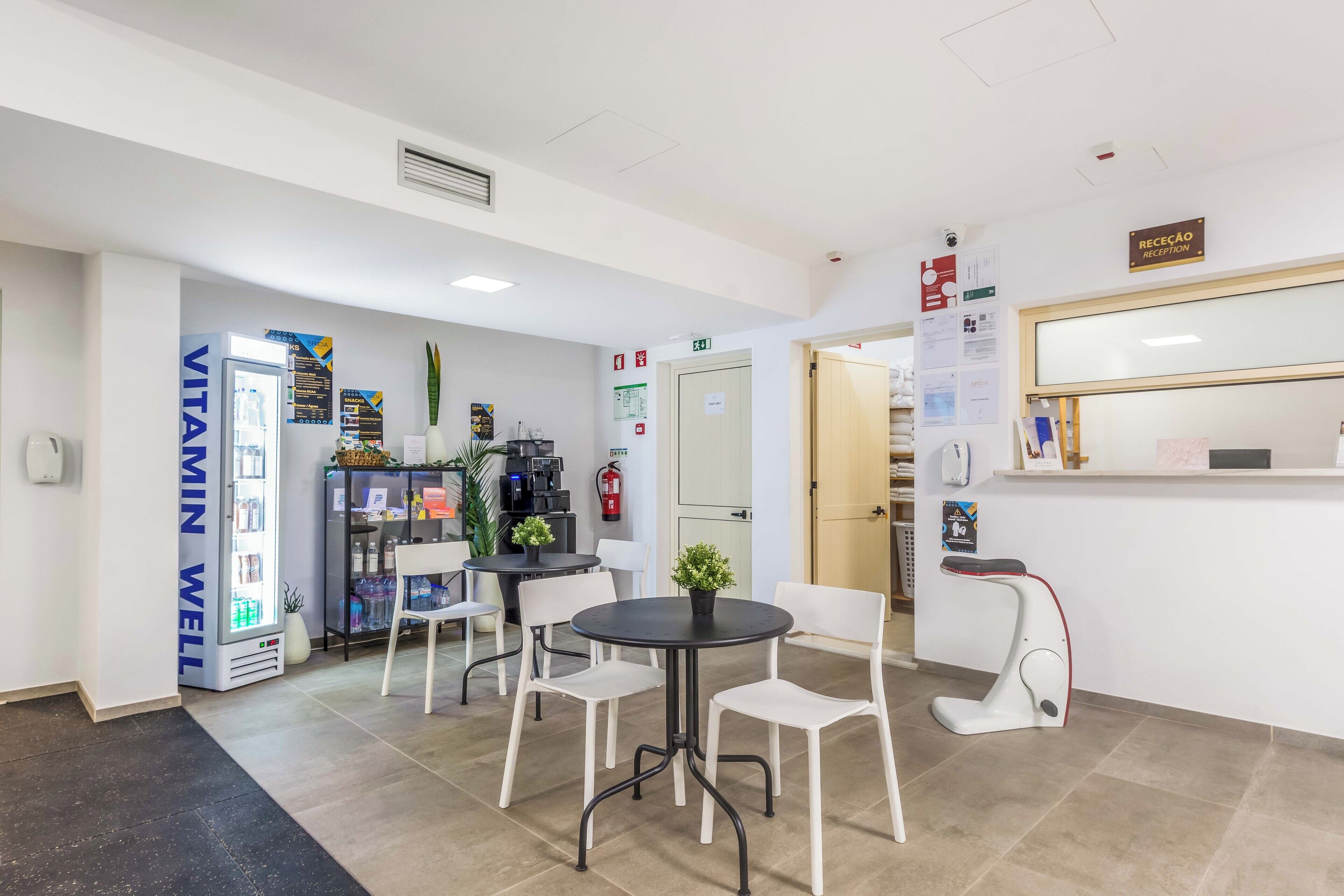 A reception area with a reception desk, a waiting area with chairs and a table, a refrigerator labeled 'VITAMIN WELL', and a coffee machine.