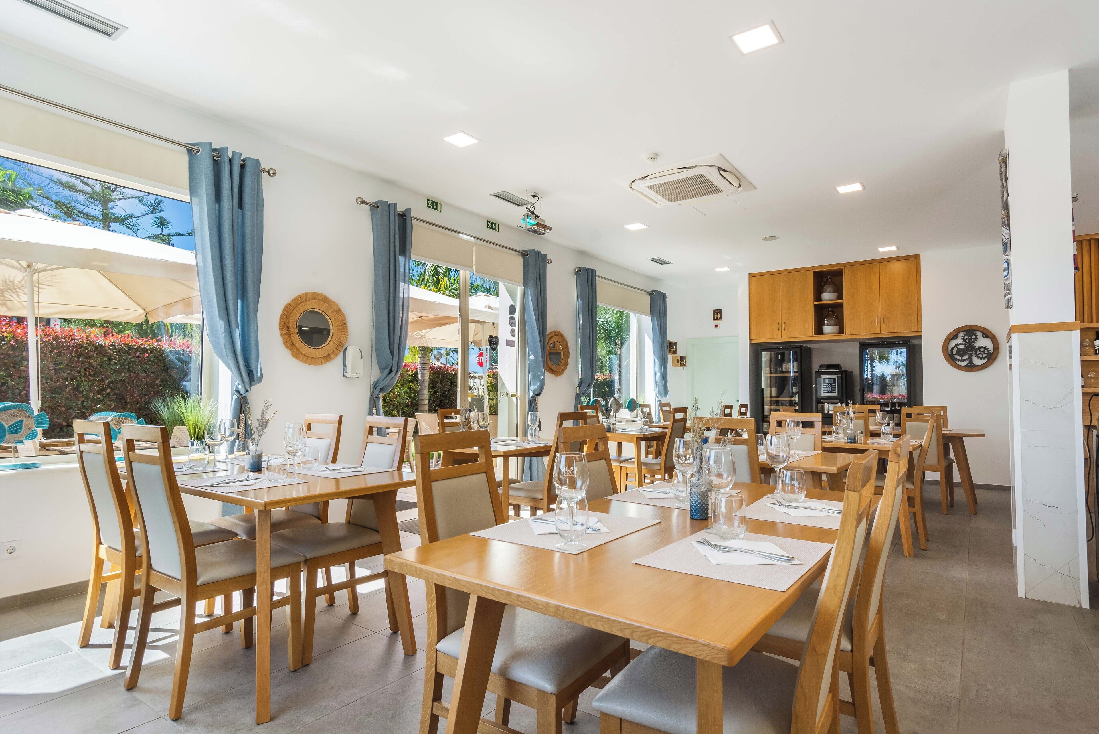 A restaurant with multiple wooden tables and chairs, large windows with curtains, and a kitchen area visible in the background.