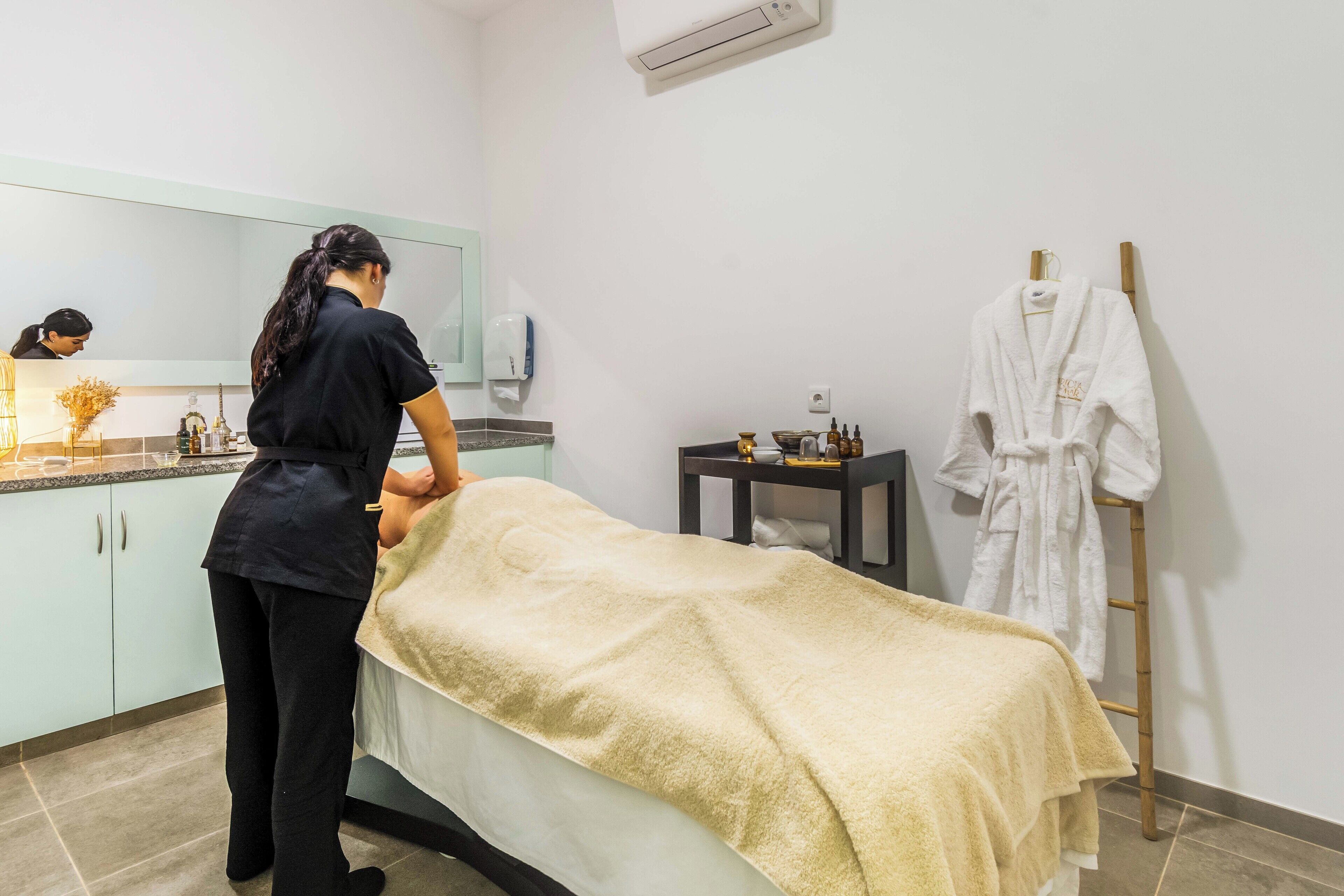 A massage room with a massage table, a towel, a massage table, a wooden side table, and a white robe on a rack.