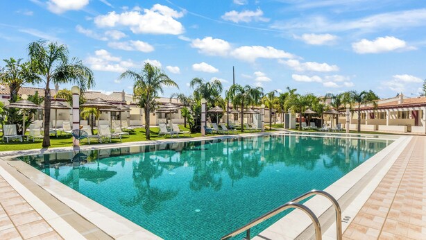 A swimming pool with lounge chairs and umbrellas, surrounded by palm trees and buildings with tiled roofs. - Hotel Rural Brícia du Mar (Lagoa)