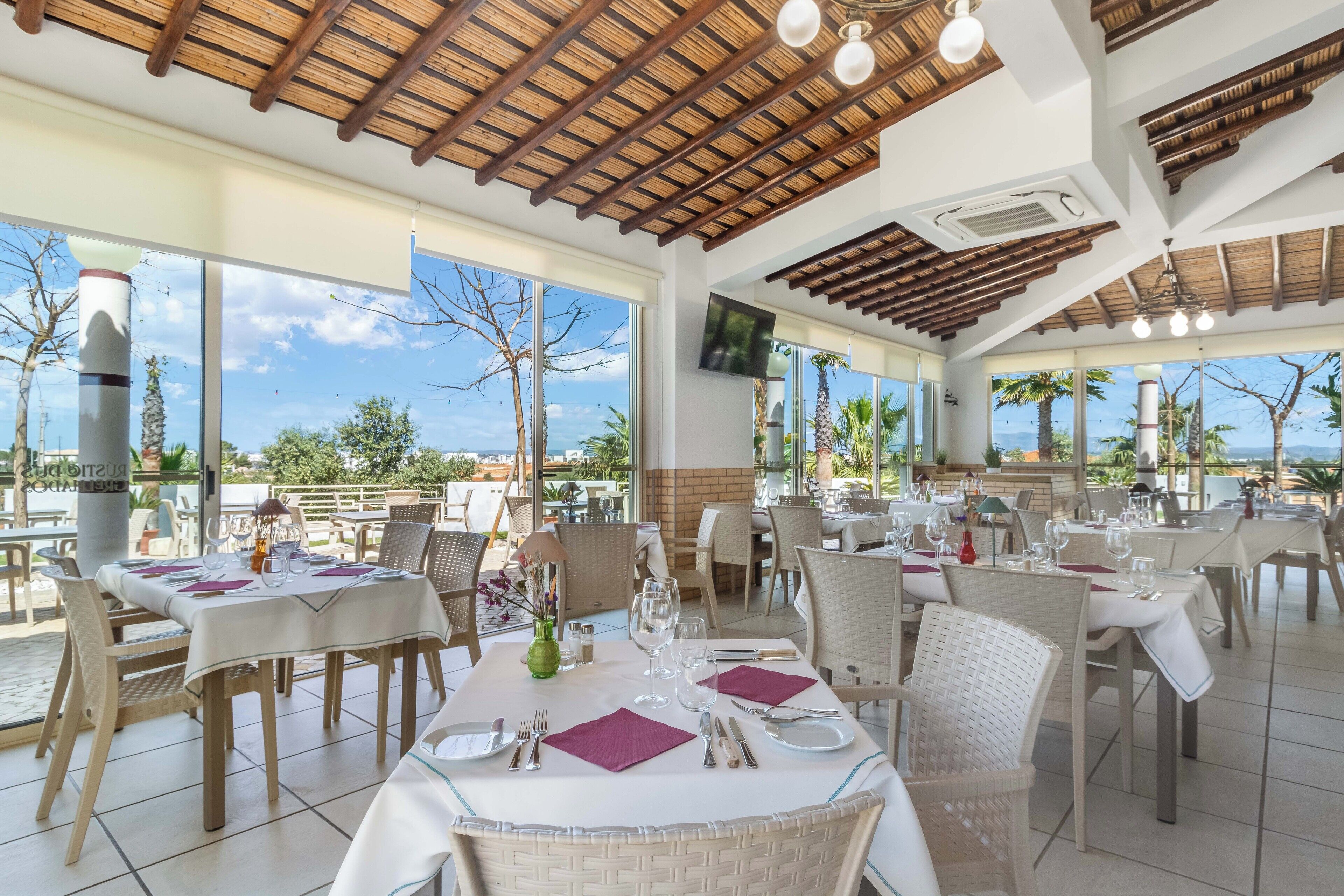 A restaurant with outdoor seating, wooden ceiling beams, and large windows offering a view of palm trees and the ocean.