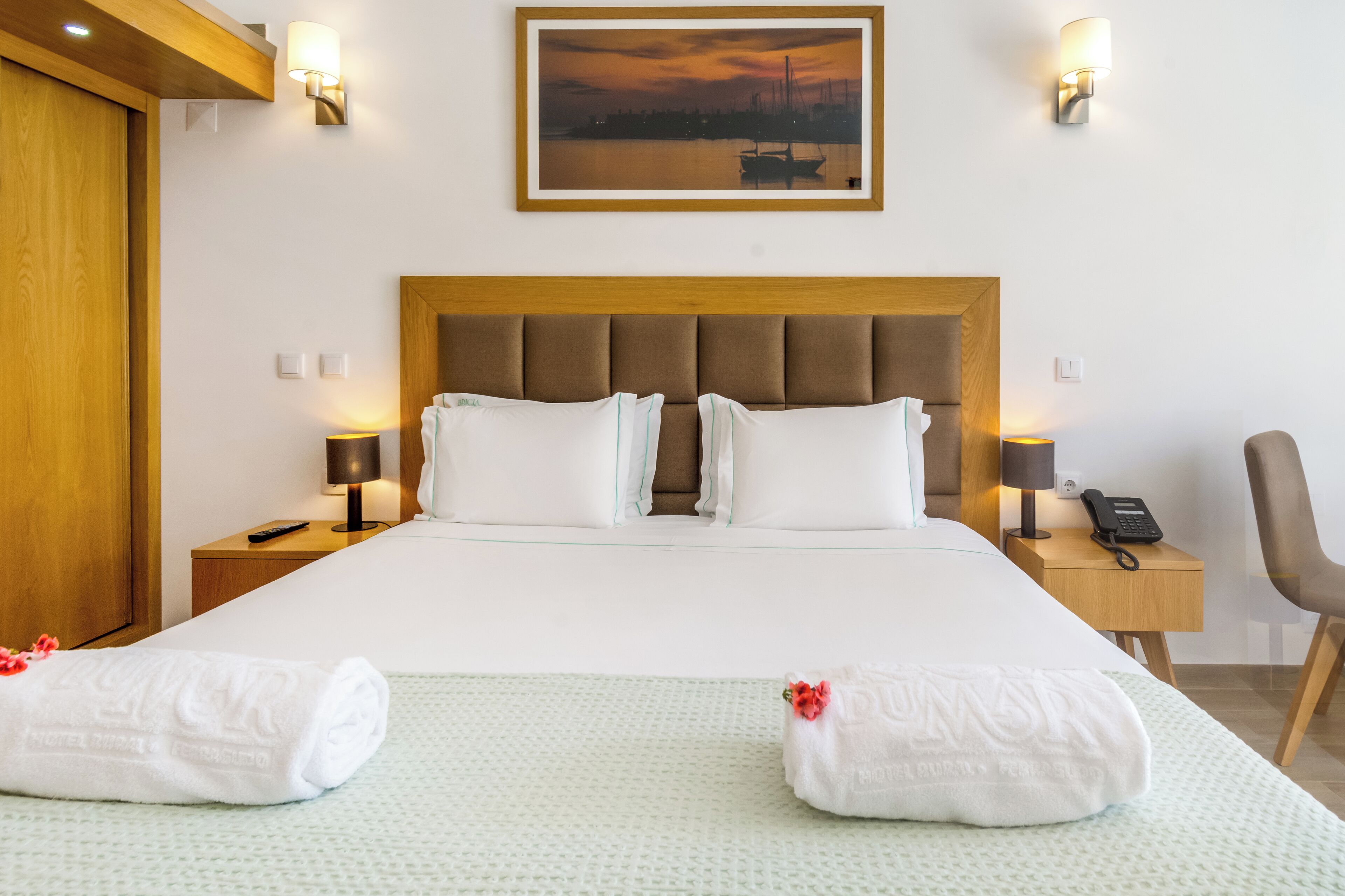 A hotel room with a large bed, two bedside tables with lamps, a chair, a painting on the wall, and a wooden door.