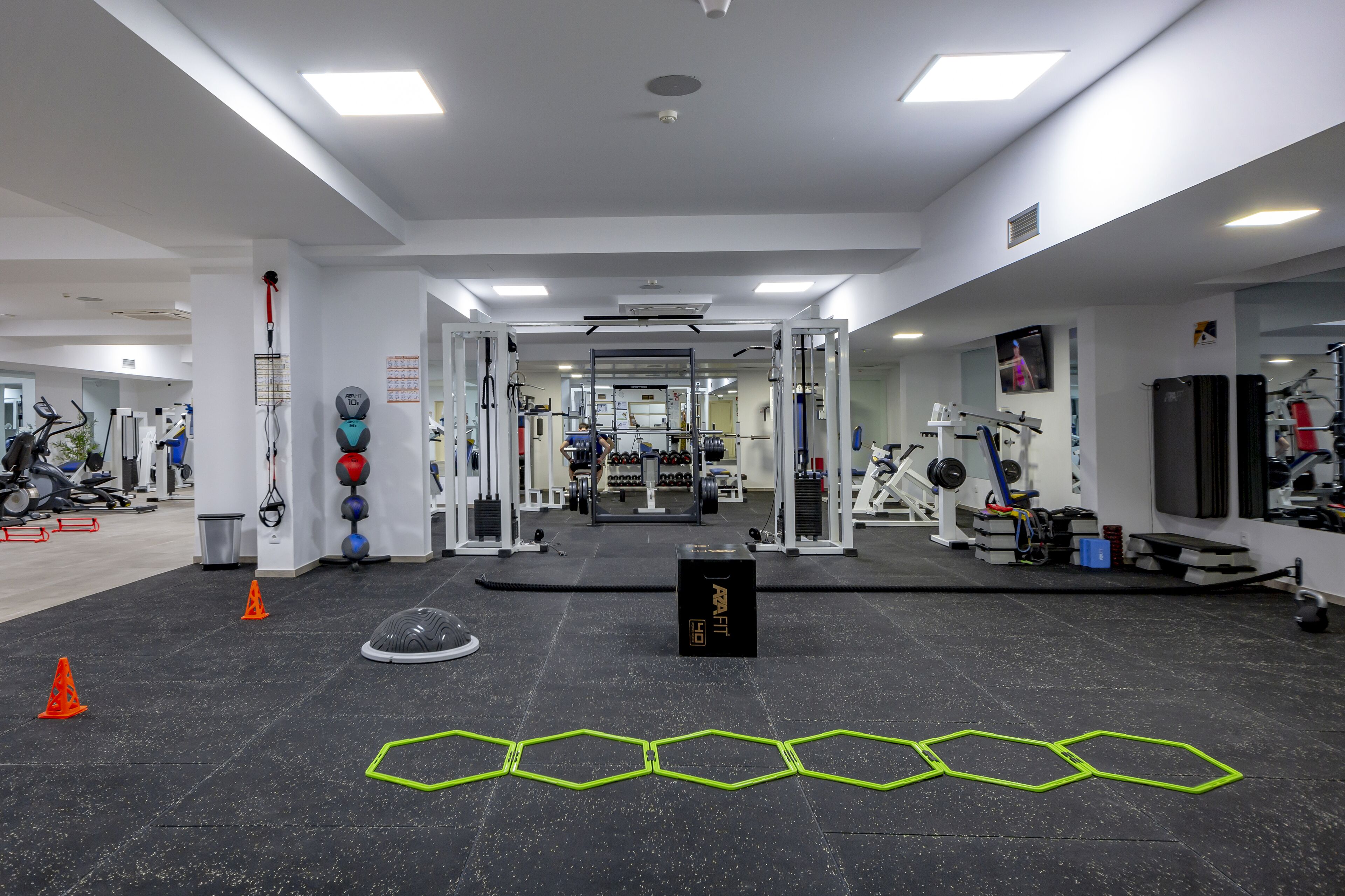Une salle de sport équipée de divers appareils d’exercice, dotée d’un mur recouvert de miroirs et d’un tapis noir orné de motifs hexagonaux sur le sol.