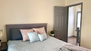 Un dormitorio con cama, almohadas, una mesita de noche, una planta en maceta y una puerta.
