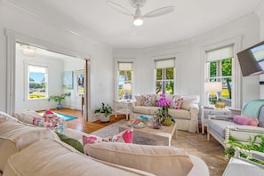 A spacious living room with a large sofa, a coffee table, and a television. There are multiple windows providing natural light and views of the outdoors. - Stella Blu - Chesapeake Properties (Cape Charles)