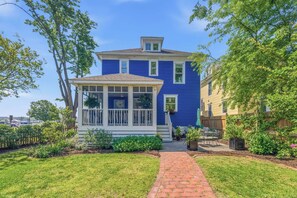 A two-story house with a blue exterior, a covered porch, and a brick pathway leading to the entrance. - Stella Blu - Chesapeake Properties (Cape Charles)