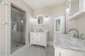 A modern bathroom with a glass shower enclosure, a double vanity with a marble countertop, and a large mirror above the sink. - Stella Blu - Chesapeake Properties (Cape Charles)