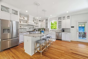 A modern kitchen with white cabinetry, stainless steel appliances, and a central island with bar stools. - Stella Blu - Chesapeake Properties (Cape Charles)