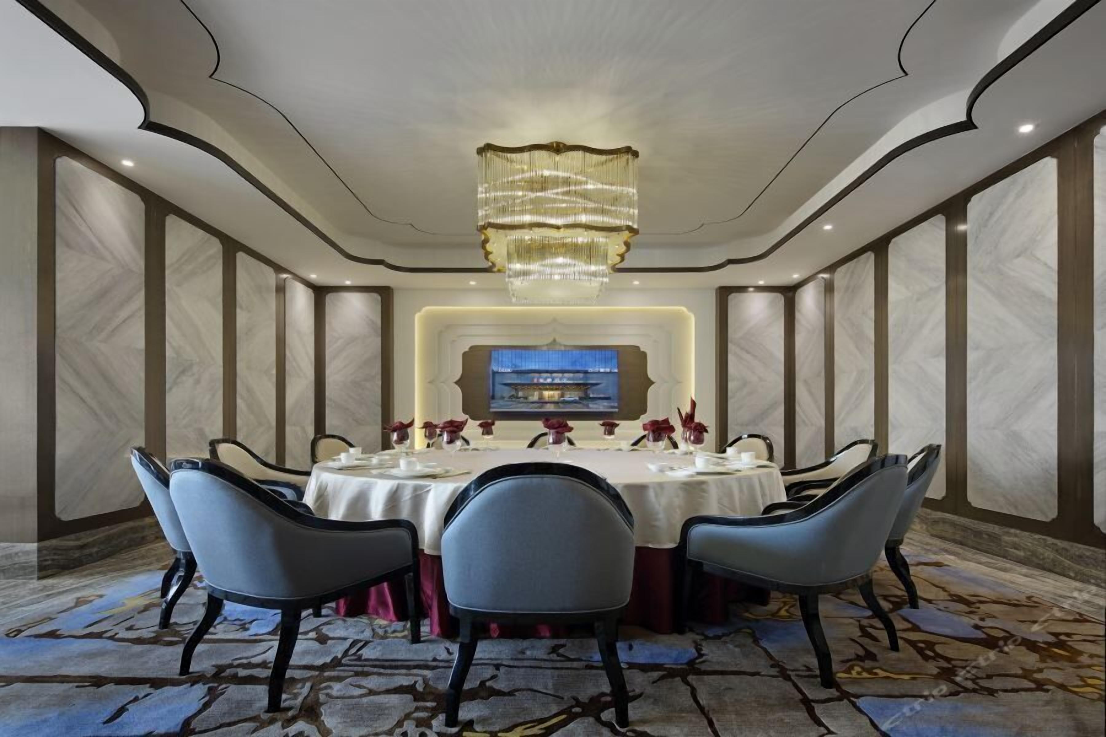A formal dining room with a round table, six chairs, a chandelier, and a large screen displaying a blue background.