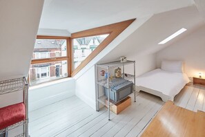 Un pequeño y luminoso dormitorio en el ático, con techo inclinado, una estantería metálica, una cama y ventana con vistas a una zona residencial.