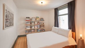Une chambre à coucher avec un grand lit, une bibliothèque, un tableau encadré et une fenêtre avec des rideaux.