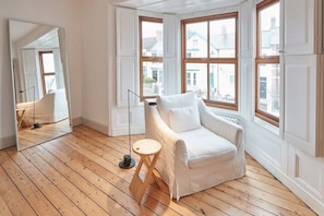 A room with a white armchair, a small wooden stool, and a floor lamp. There are large windows with wooden frames.