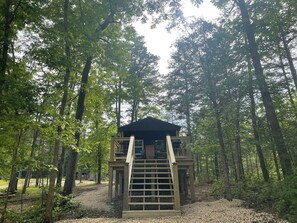 A wooden cabin with a staircase leading up to a covered entrance, surrounded by a forest.