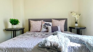 A neatly made bed with a patterned bedspread, multiple pillows, and a fluffy throw blanket. There are two nightstands with potted plants and a small table beside the bed.