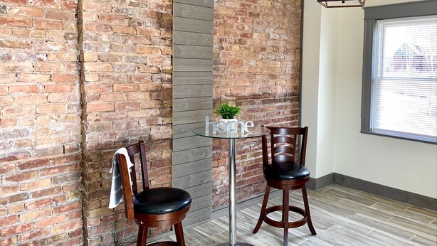 A room with a brick wall, a glass table with two wooden chairs, a hanging light fixture, and a window with blinds.