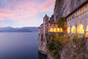 Un bâtiment historique doté d’une tour et de fenêtres en arc de cercle, situé sur une falaise surplombant un lac paisible, avec des montagnes en arrière-plan.