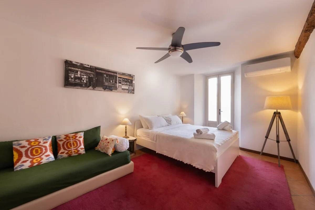 Camera d'albergo con un letto, un divano verde, una ventola a soffitto, una lampada e un quadro appeso al muro.