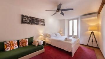 Camera d'albergo con un letto, un divano verde, una ventola a soffitto, una lampada e un quadro appeso al muro.
