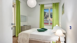 A bedroom with a bed, a bedside table with a lamp, a window with curtains, and a view of buildings outside.