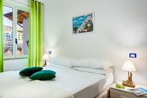 Una cama bien hecha con almohadas verdes, una mesita de noche con lámpara, y un cuadro con una escena costera en la pared.