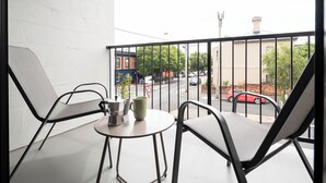 Un balcón con dos sillas, una mesa pequeña y vista a una calle con coches y edificios.