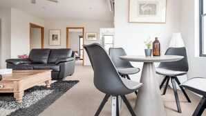 Una sala de estar moderna con un sofá de cuero negro, una mesa redonda de comedor con cuatro sillas, una mesa de centro y un amplio ventanal que proporciona luz natural.