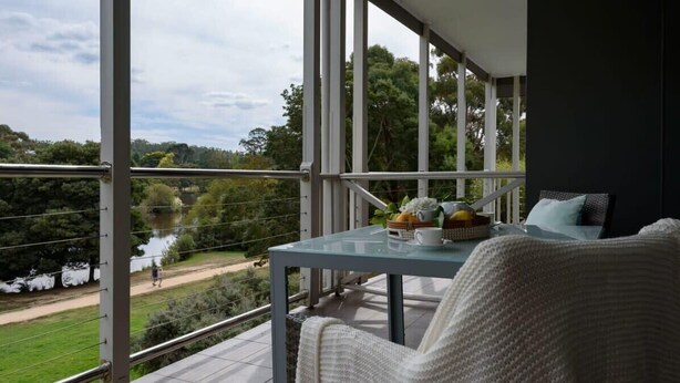 A balcony with a glass table, a wicker chair, and a view of a river and greenery. - Lakeside Suite 1 + Lakeside Retreat + Spa Bath (Daylesford)