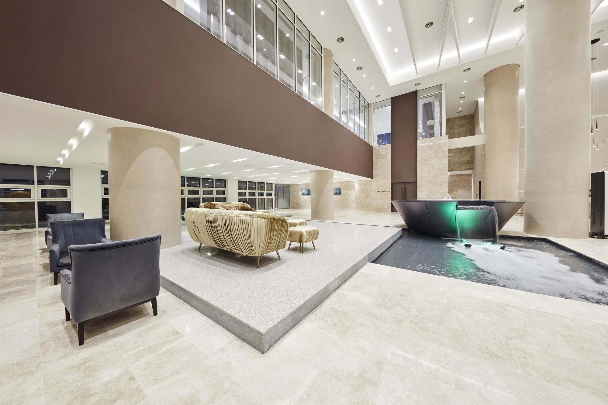 A spacious lobby with a modern design, featuring a seating area with sofas and chairs, a water feature, and a high ceiling with natural light.