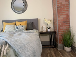 A bedroom with a bed, a bedside table with a framed quote, a potted plant, and a mirror on the wall.