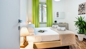 A modern bedroom with a bed, a bedside table with a lamp, a window with green curtains, a potted plant, and artwork on the wall. - Loving Times Apartment (Sorrento)
