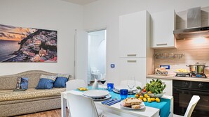 A modern kitchen with white cabinetry, a dining area with a table set for two, and a living room with a patterned sofa. - Loving Times Apartment (Sorrento)