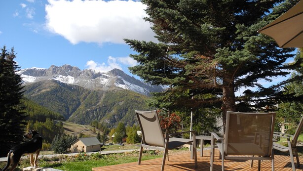 Una terraza de madera con mobiliario exterior, un gran árbol de hoja perenne y una vista de la montaña al fondo.