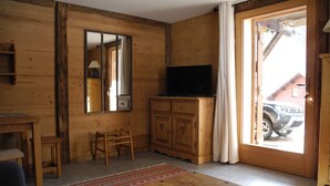 Una habitación con paneles de madera, un televisor sobre un mueble bajo, una silla, una mesita pequeña y un ventanal con cortinas.