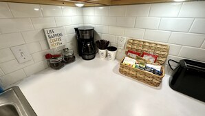 Une cuisine avec des carreaux blancs, une machine à café, des bocaux et un panier rempli d’en-cas.