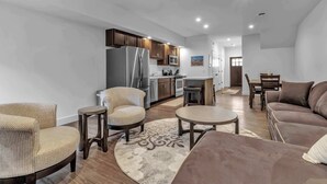 Una sala de estar moderna con sofá, sillones, una mesa de centro y una zona de cocina con refrigerador, microondas y armarios.