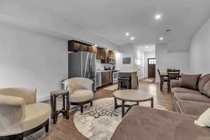 Una sala de estar moderna con sofá, sillones, una mesa de centro y una zona de cocina con refrigerador, microondas y armarios.