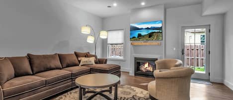 A living room with a sofa, armchair, coffee table, fireplace, TV, and a view of the outdoors.