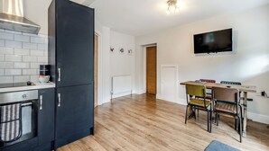 Una cocina moderna con un refrigerador negro, un horno empotrado y una cafetera. Hay un comedor con una mesa y sillas de madera, y un televisor montado en la pared.