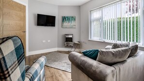 Una sala de estar moderna con un sofá gris, un sillón escocés, un televisor de pantalla plana, una chimenea y un ventanal grande con persianas.
