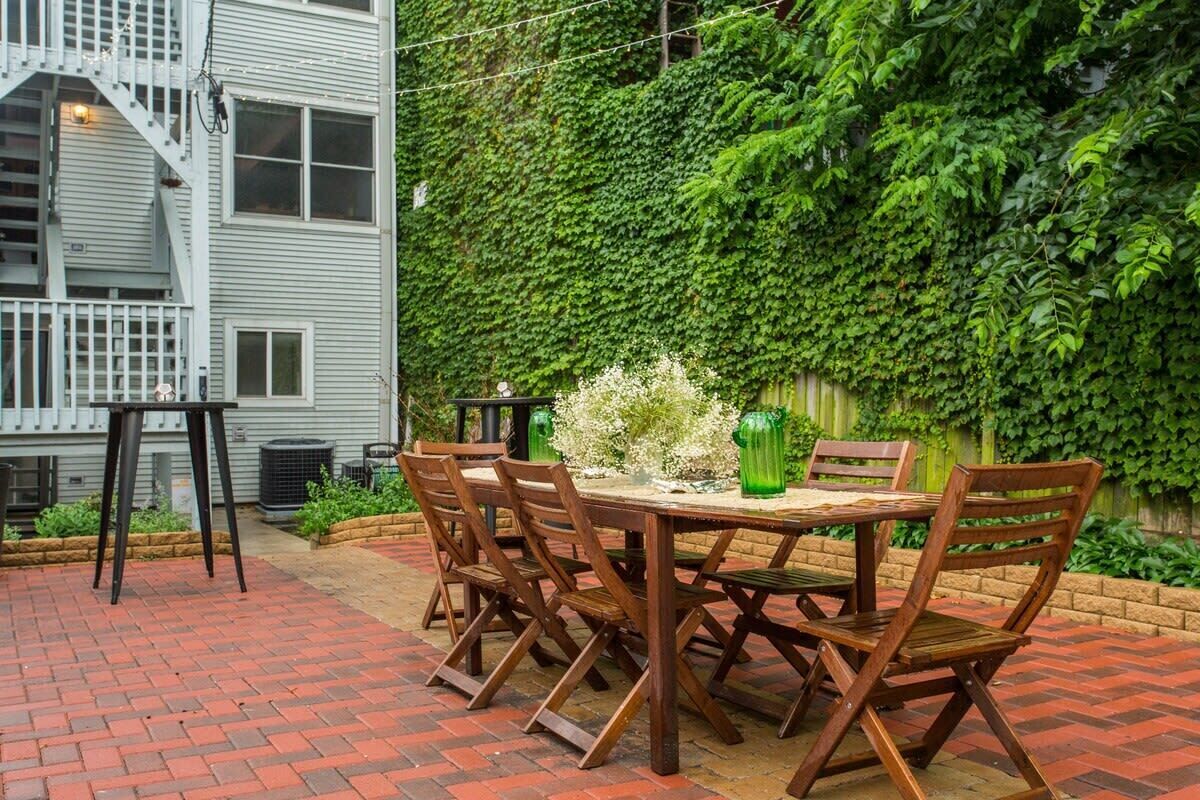 Un patio con una mesa y sillas de madera, rodeado de un suelo de ladrillo y una pared verde cubierta de hiedra.
