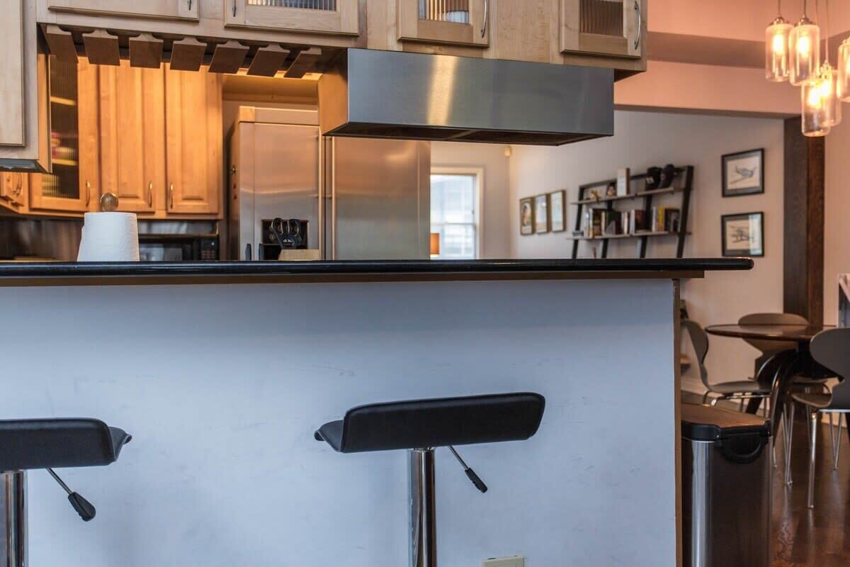 A modern kitchen with wooden cabinets, a stainless steel range hood, and a bar area with two black stools.