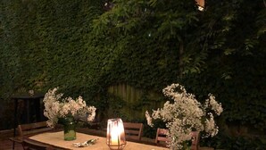 Un área de comedor al aire libre con una mesa preparada para cuatro personas, rodeada de sillas. Hay dos jarrones verdes con flores blancas sobre la mesa, y un farol encendido en el centro. Guirnaldas de luces decoran el espacio sobre la mesa.