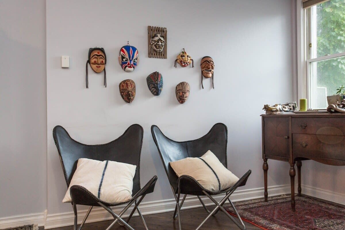A room with two black chairs with white cushions, a wooden cabinet, and a wall adorned with various masks.