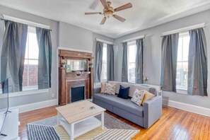 Una sala de estar con chimenea, sofá gris, piso de madera y ventanas grandes con cortinas.