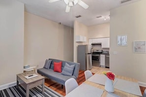 Un appartement moderne avec un canapé gris, une petite table en bois, un coin repas avec une table et des chaises blanches, et une cuisine en arrière-plan.