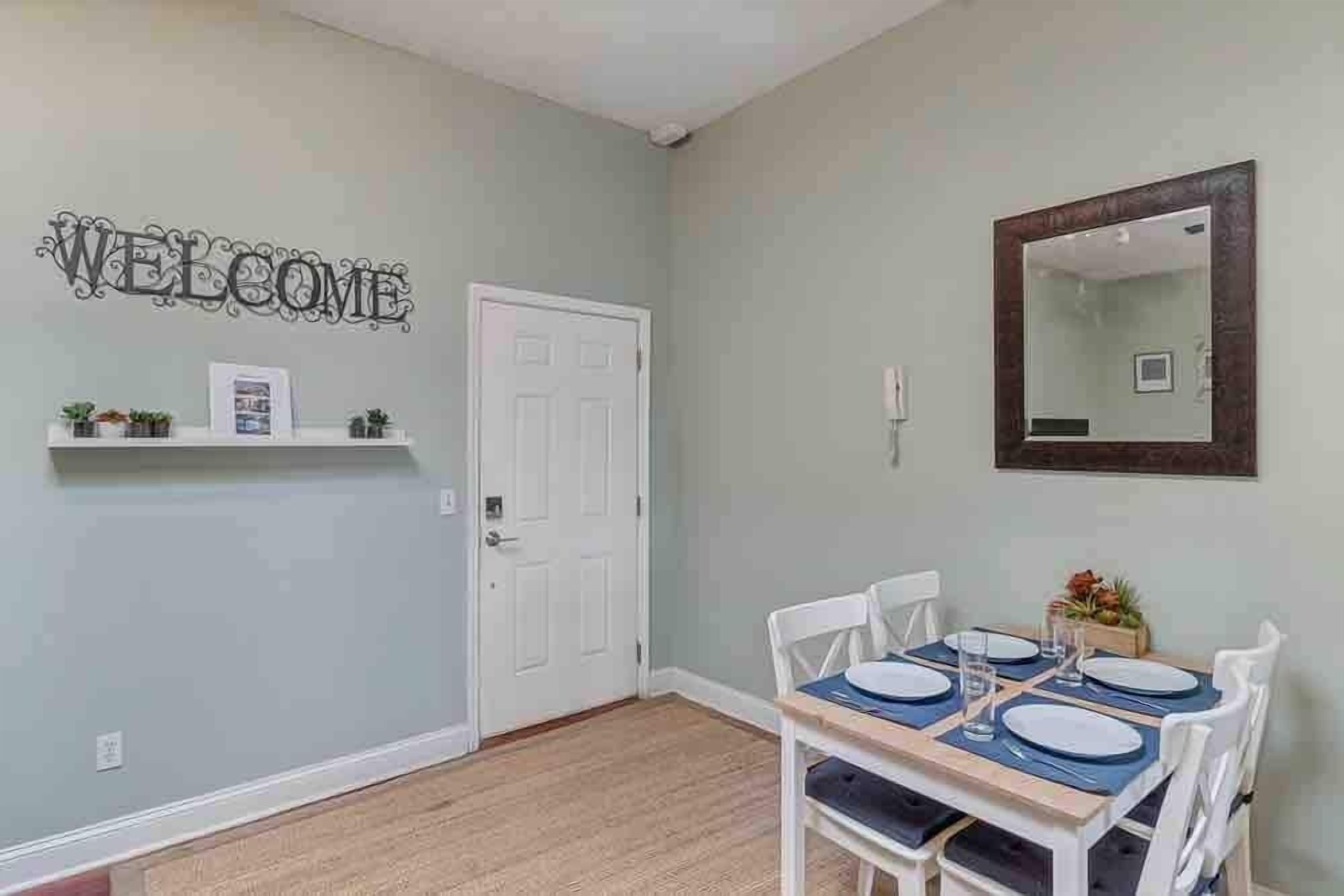 A dining area with a 'WELCOME' sign, a white door, a mirror, and a table set for four.