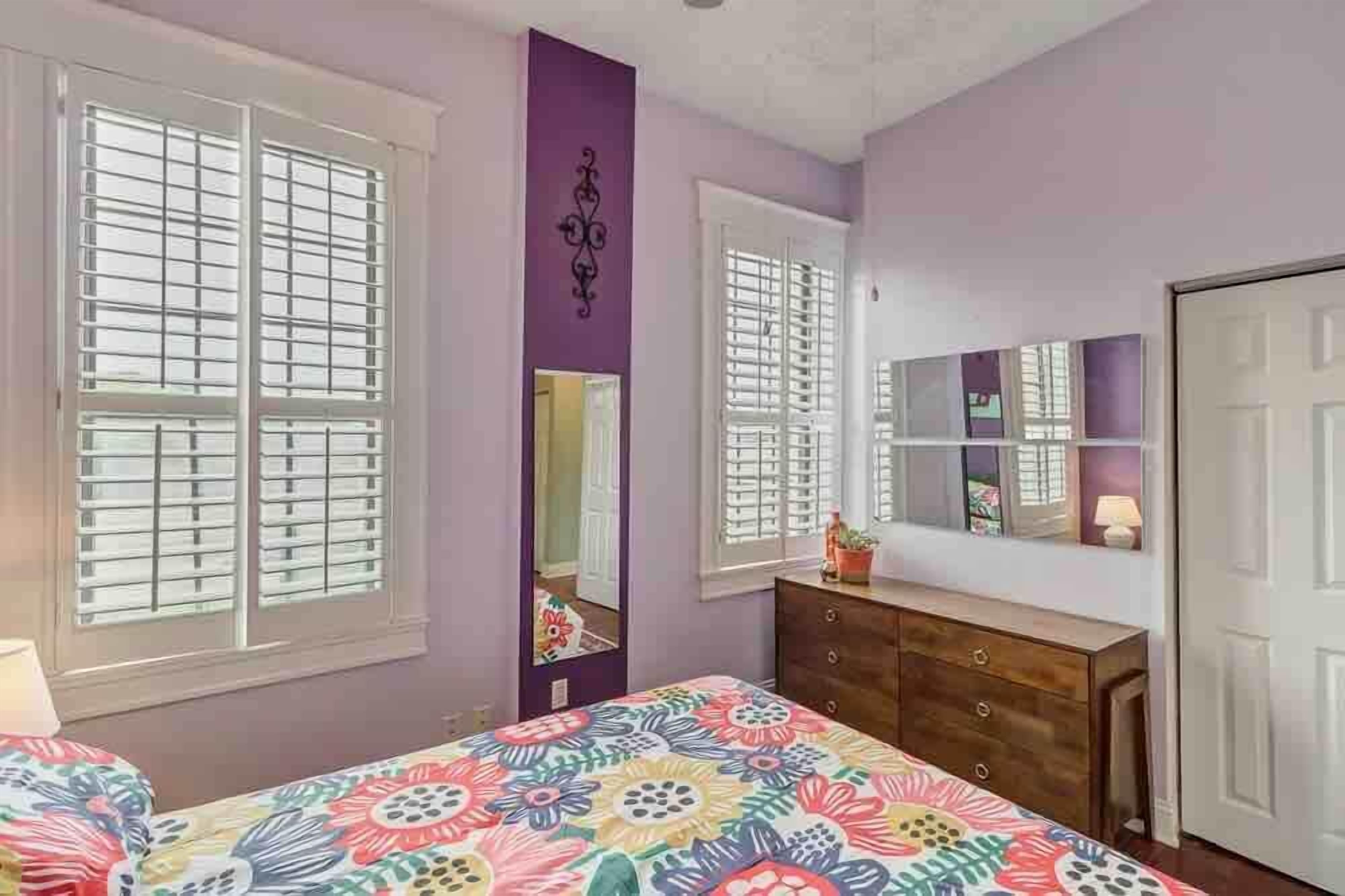 A bedroom with two windows, a dresser, a mirror, and a bed with a colorful floral bedspread.