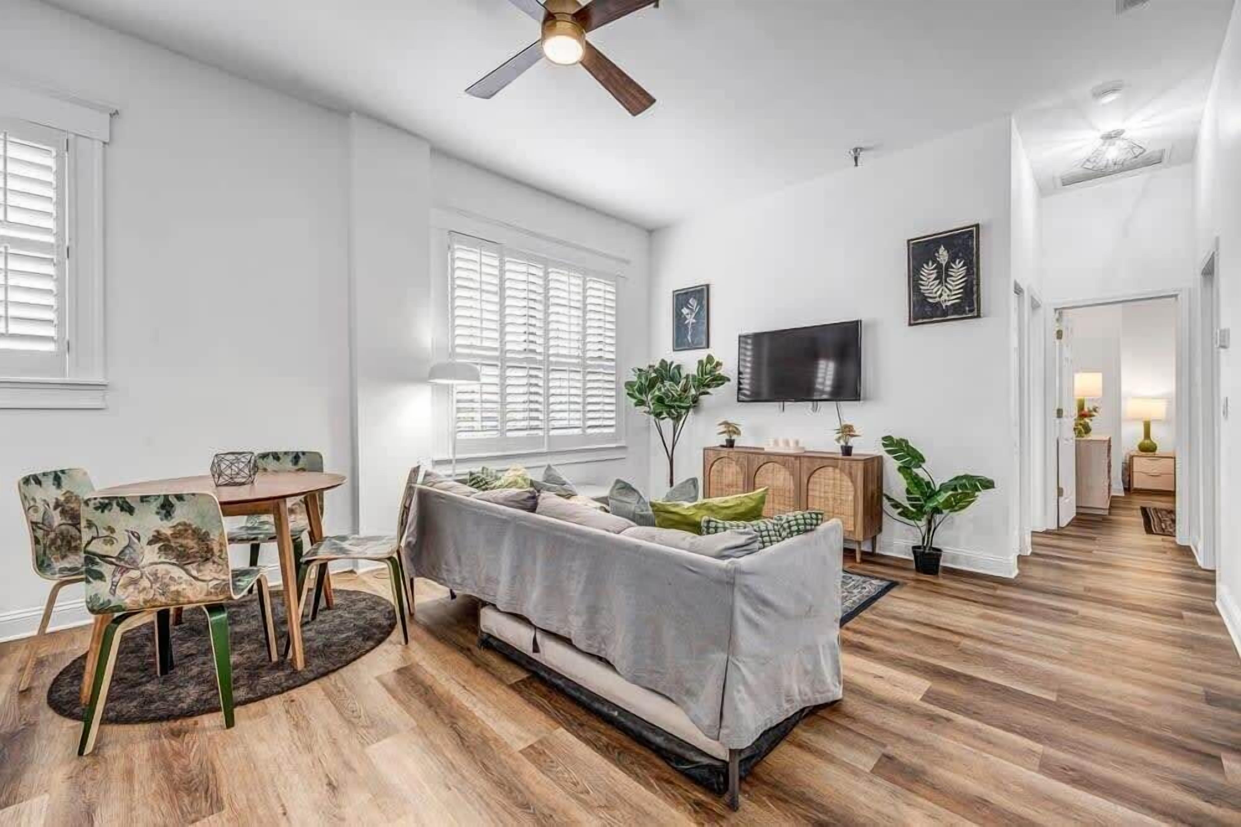 Un vaste salon comprenant un canapé gris, une table à manger en bois et des chaises, un ventilateur de plafond et un téléviseur fixé au mur.