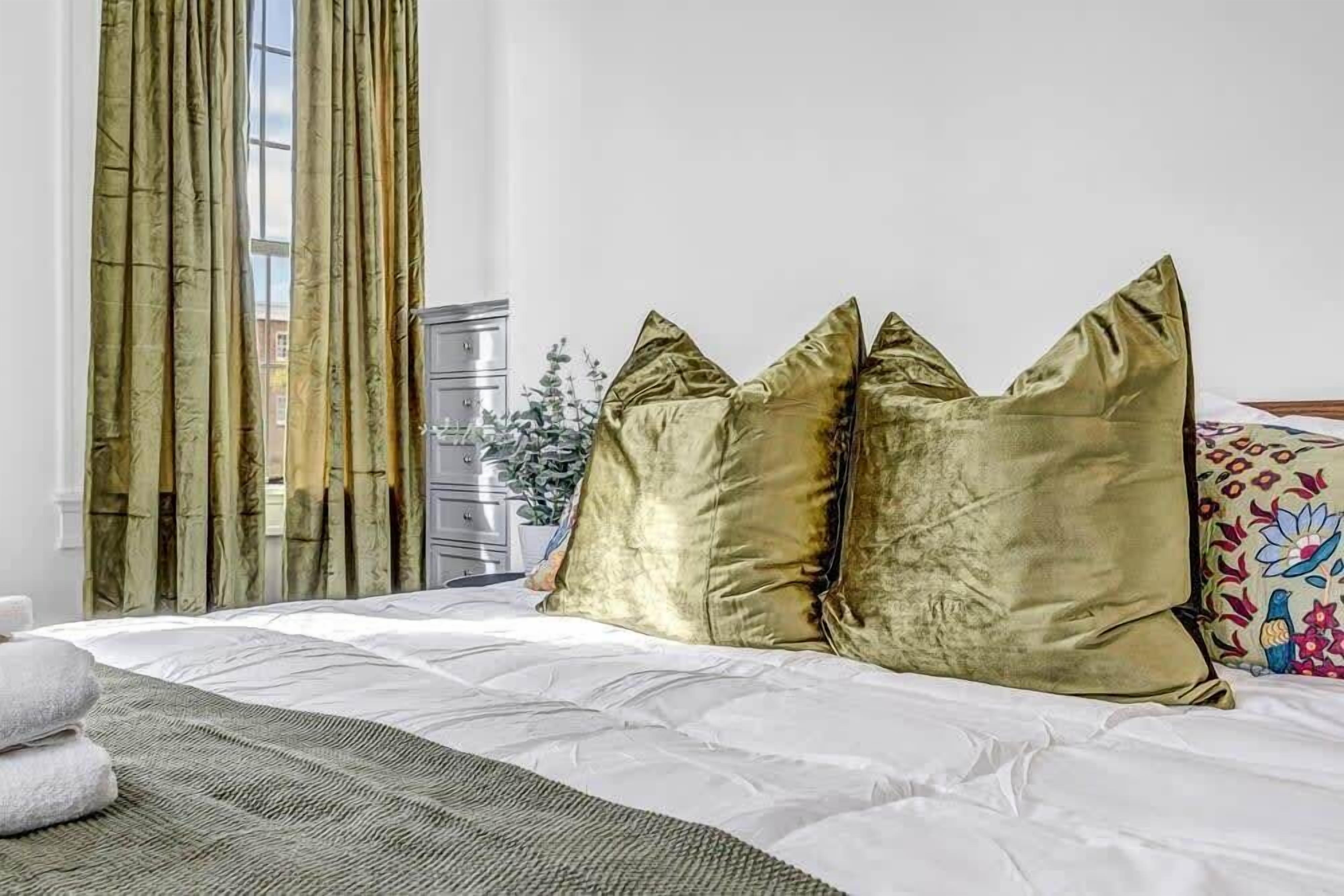 Un lit bien fait, avec une couette blanche et un plaid vert, agrémenté de trois coussins dorés.