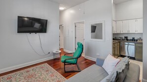 A modern living room with a flat-screen TV, a green chair, a sofa, and a kitchen area with stainless steel appliances.